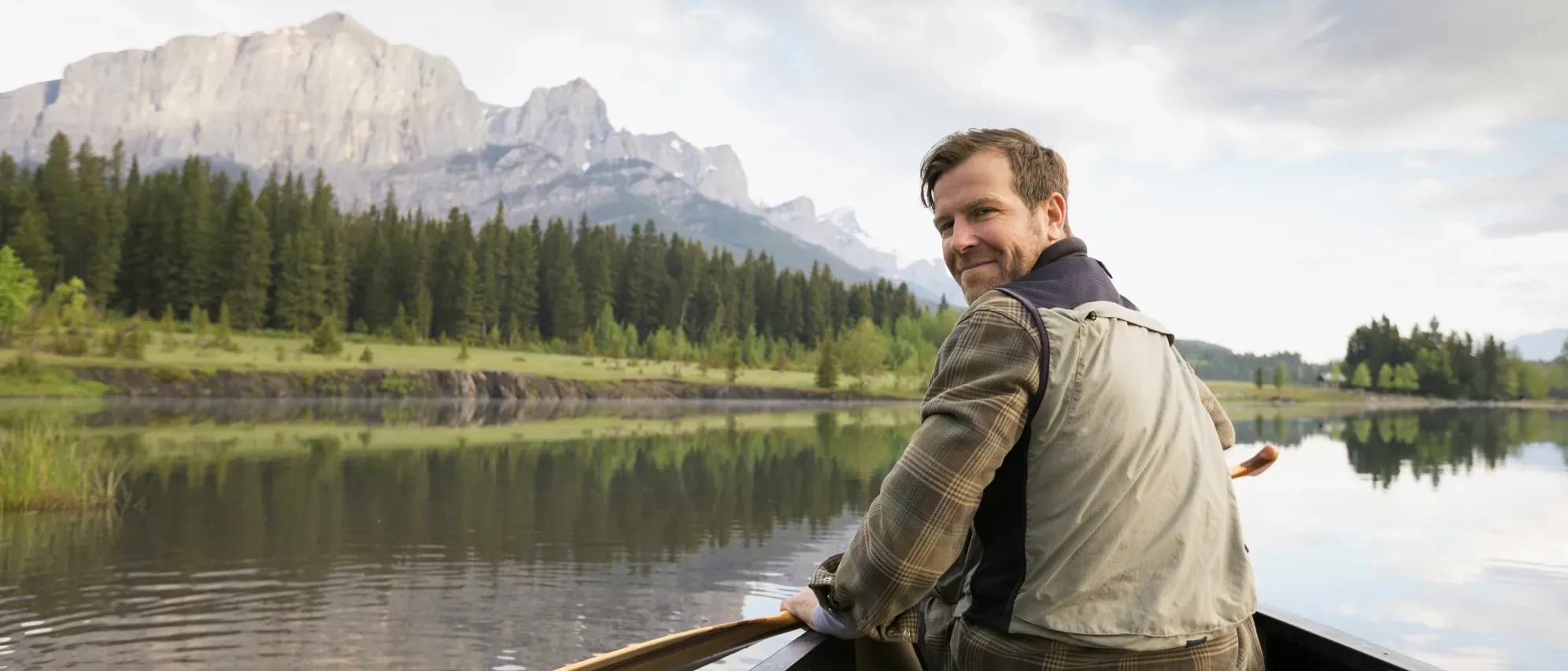 Un homme souriant est assis dans un canoë sur une rivière. Des montagnes se dressent en arrière-plan