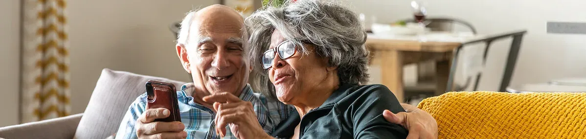 deux personnes âgées souriantes regardent leur mobile dans leur canapé