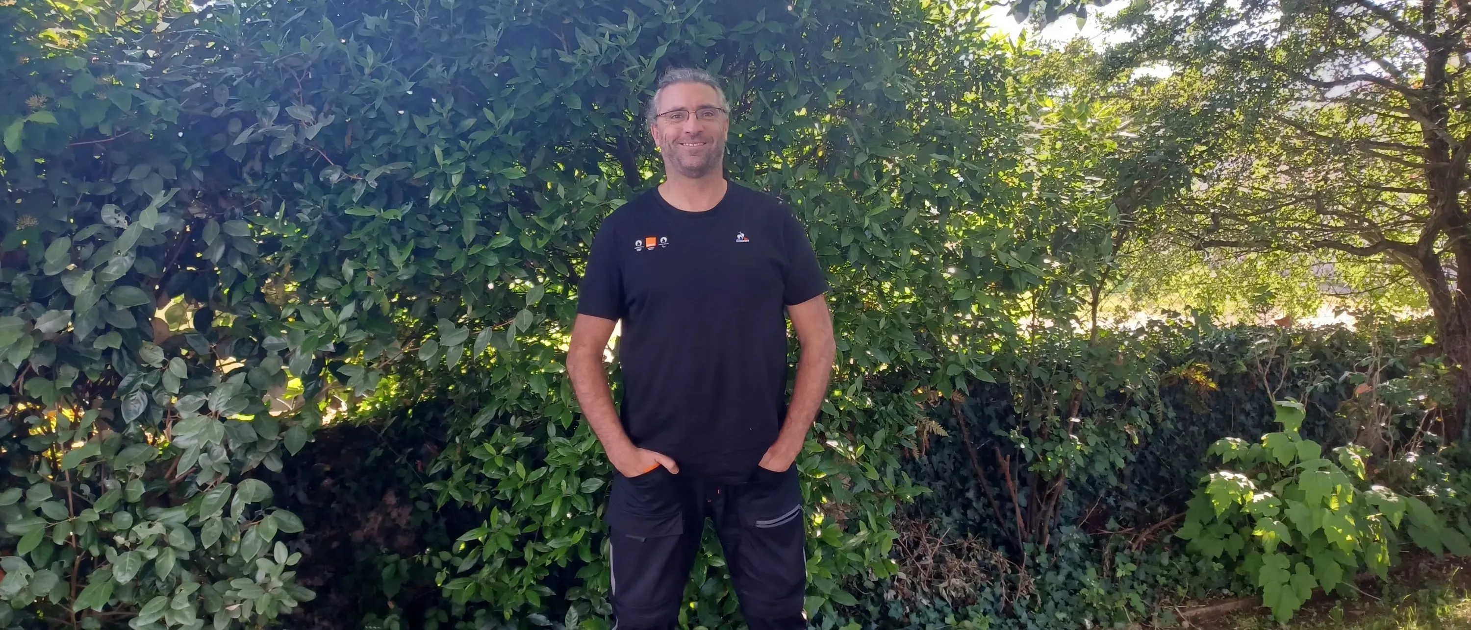 Cédric souriant devant un buisson